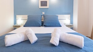 Ein ordentlich bezogenes Bett mit zwei Kissen und zwei aufgerollten Handtüchern, ein Nachttisch mit Lampe sowie ein an der Wand befestigter Bilderrahmen.