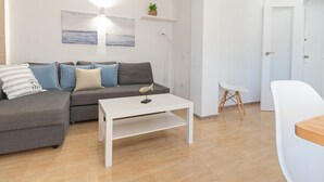 Ein modernes Wohnzimmer mit einem grauen Sofa, einem weißen Couchtisch und einem kleinen Beistelltisch. An der Wand hängt ein Gemälde, und im Hintergrund ist eine Tür zu sehen.