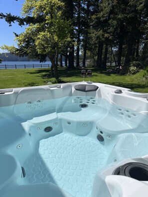 Ett bubbelbad med flera massagemunstycken, omgivet av en gräsyta och träd, med utsikt över en sjö i bakgrunden.