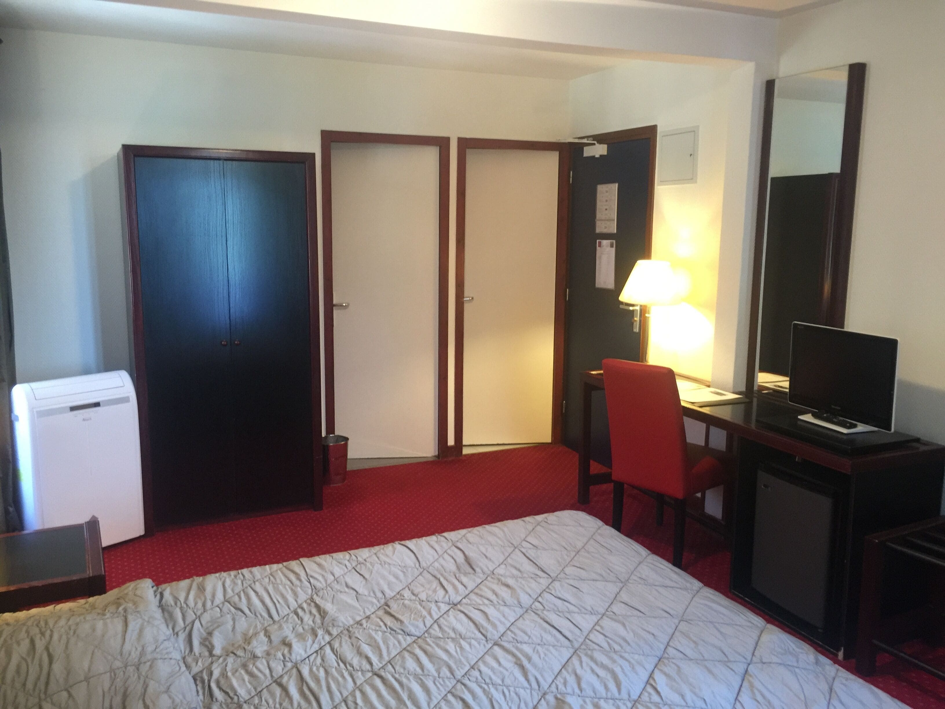 A hotel room with a bed, a desk with a chair, a television, a desk lamp, and a refrigerator.