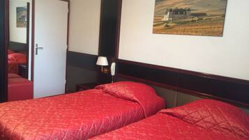 Habitación de hotel con dos camas rojas, un cuadro de un paisaje campestre, una lámpara de noche y una puerta que da a otra habitación.