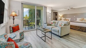 A modern hotel room with a sofa, a coffee table, a TV, a balcony with a view of palm trees, and a bed with a headboard. - 2 QUEEN BEDS | FREE WIFI |  HEATED POOLS | FREE PARKING | STEPS TO BONITA BEACH (Bonita Springs)