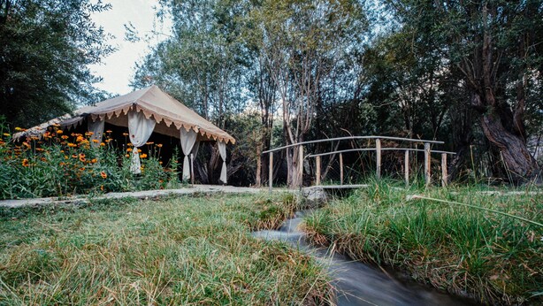 Un puente de madera rústico sobre un pequeño arroyo, una tienda de campaña con toldo y un entorno natural con árboles y hierba.