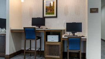 A business center with two desks, each with a computer and a chair. There is a framed picture on the wall and a sign indicating the business center location.
