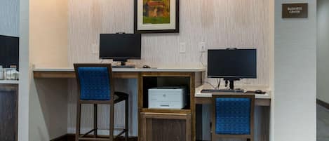 A business center with two desks, each with a computer and a chair. There is a framed picture on the wall and a sign indicating the business center location.