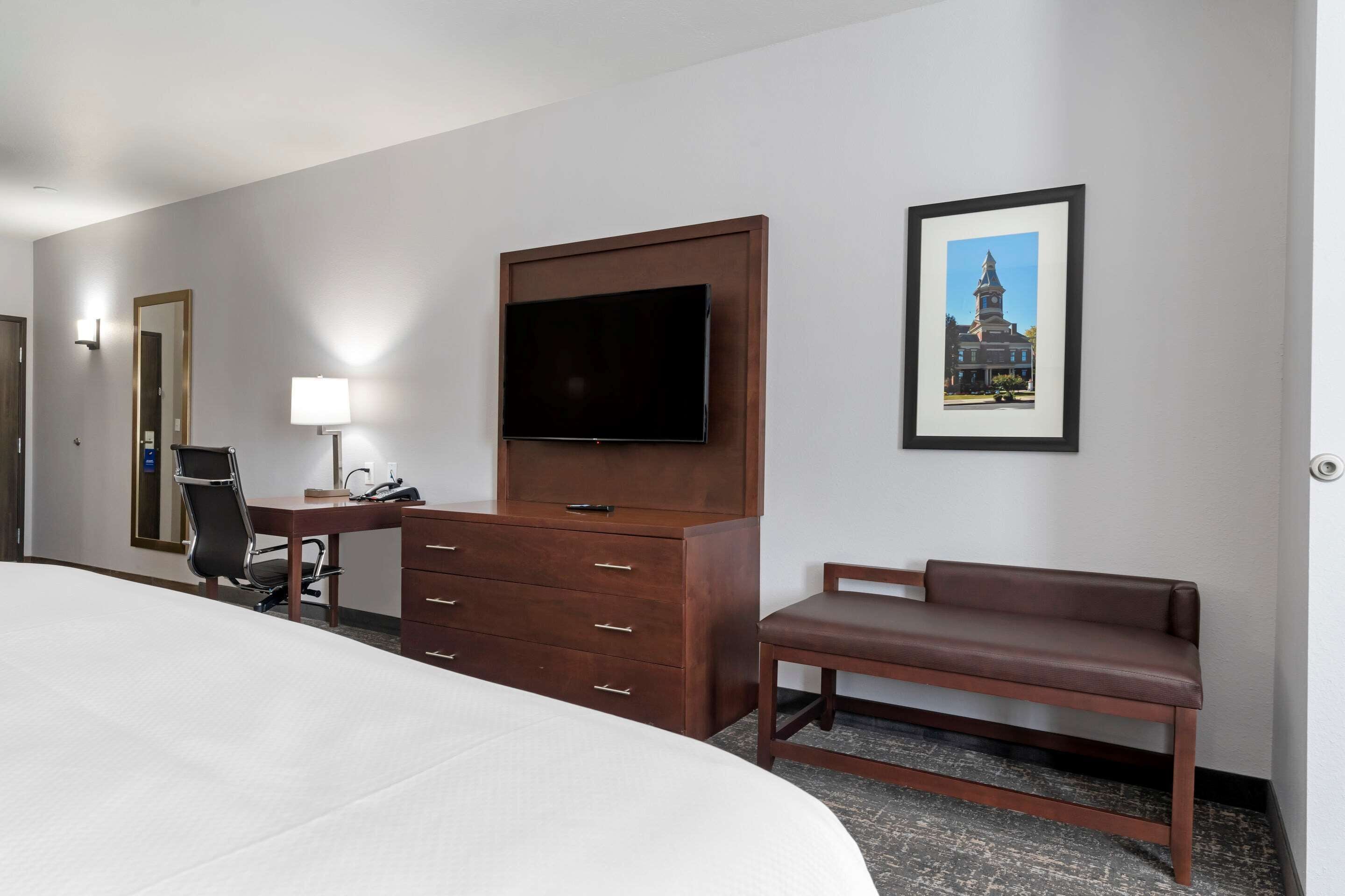 A hotel room with a large bed, a desk with a chair, a TV mounted on a wooden cabinet, a bench, and a framed picture on the wall.