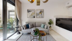 Een moderne woonkamer met een grijze bank, twee abstracte schilderijen, een televisie en een glazen deur die leidt naar een balkon met planten.
