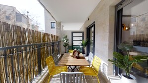 Een balkon met een houten tafel, gele stoelen en plantenpotten.