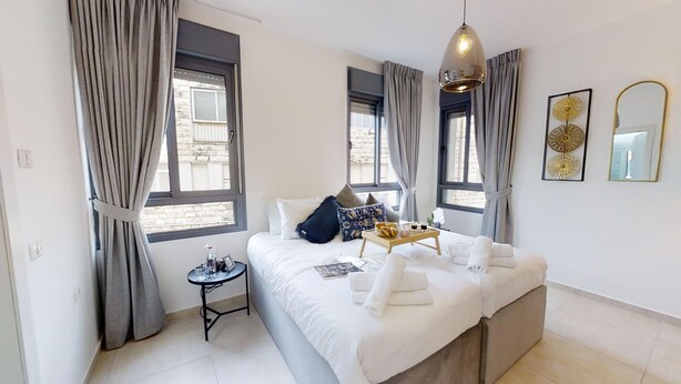 Een moderne slaapkamer met een groot bed, twee ramen met gordijnen, een nachtkastje met lamp en decoratieve muurkunst.