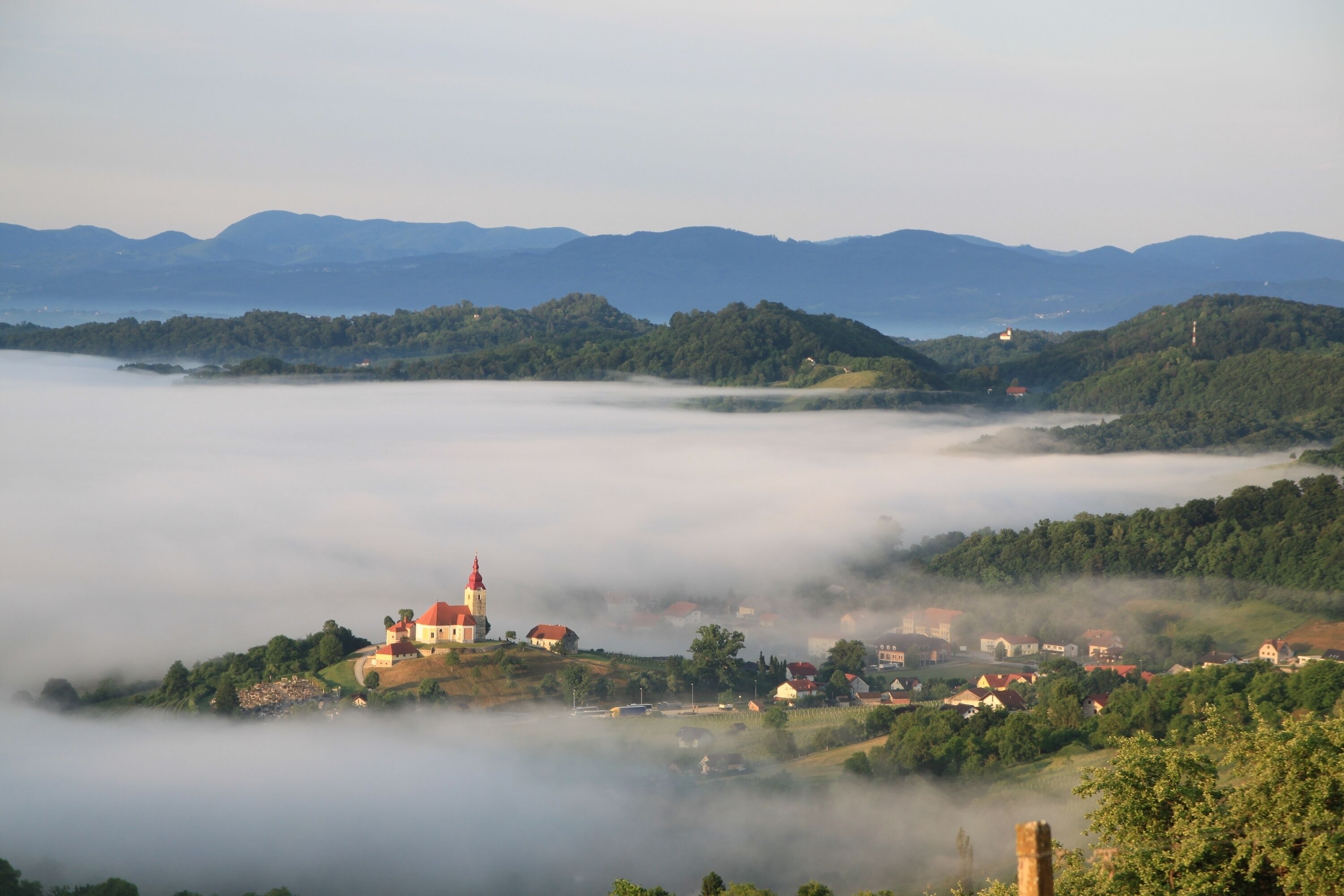 A village with a prominent church on a hill, surrounded by lush greenery and mist.