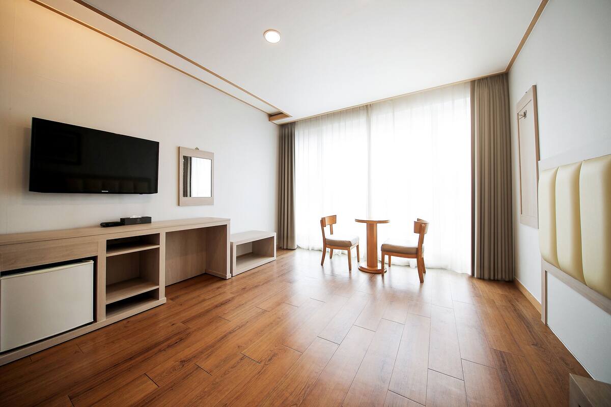 A modern hotel room with a wooden floor, a flat-screen TV mounted on the wall, a desk with a chair, and a window with curtains.