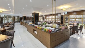 A spacious hotel lobby with a central buffet area, multiple seating arrangements, and large windows providing natural light.