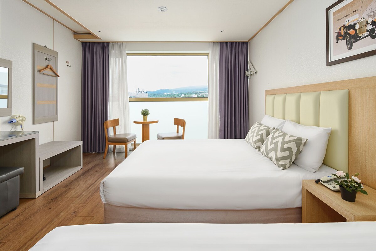A hotel room with a large bed, a desk with a chair, a small table with two chairs, and a view of the city through the window.