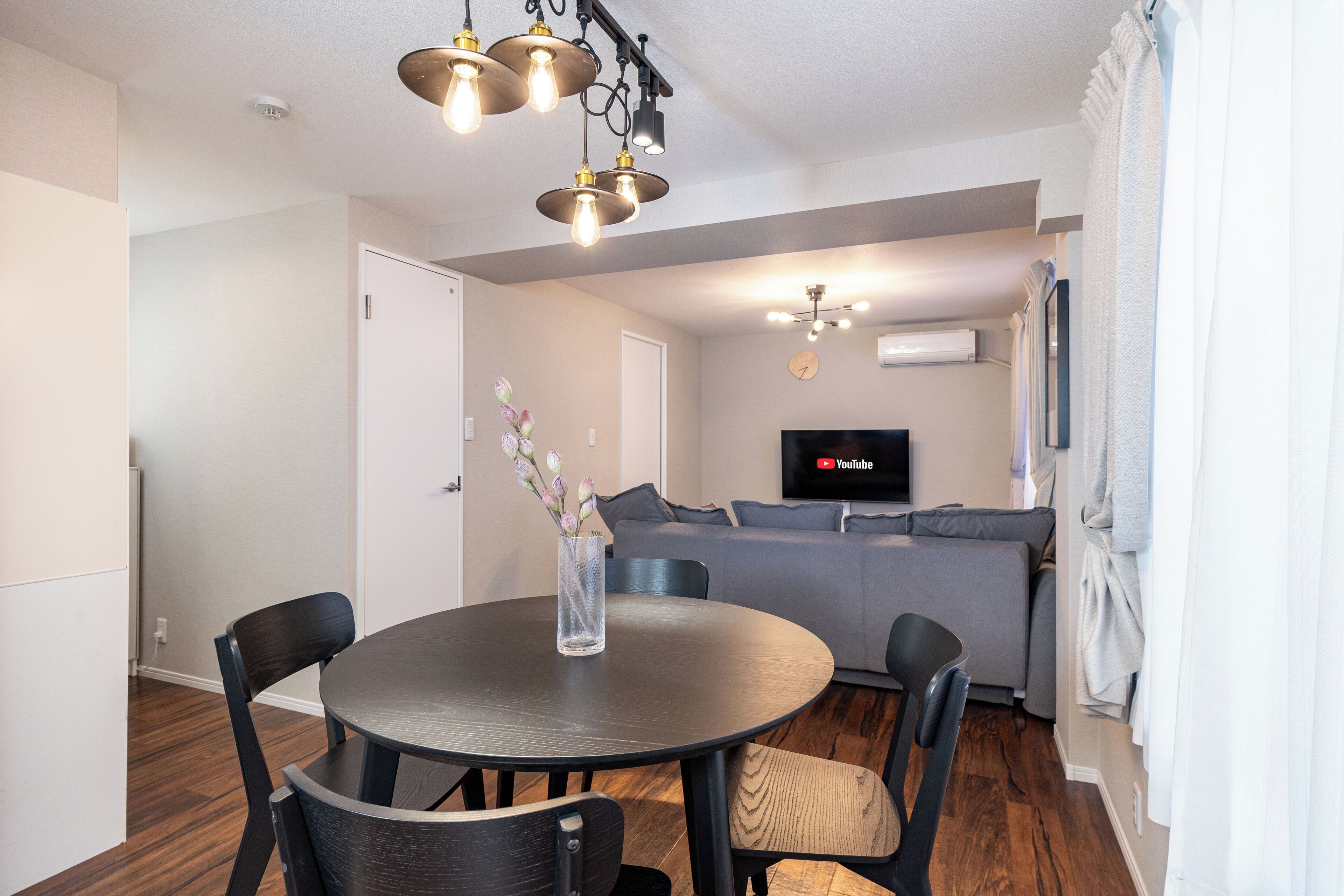 A modern living room with a grey sofa, a round dining table with chairs, a TV mounted on the wall, and a decorative vase on the table.