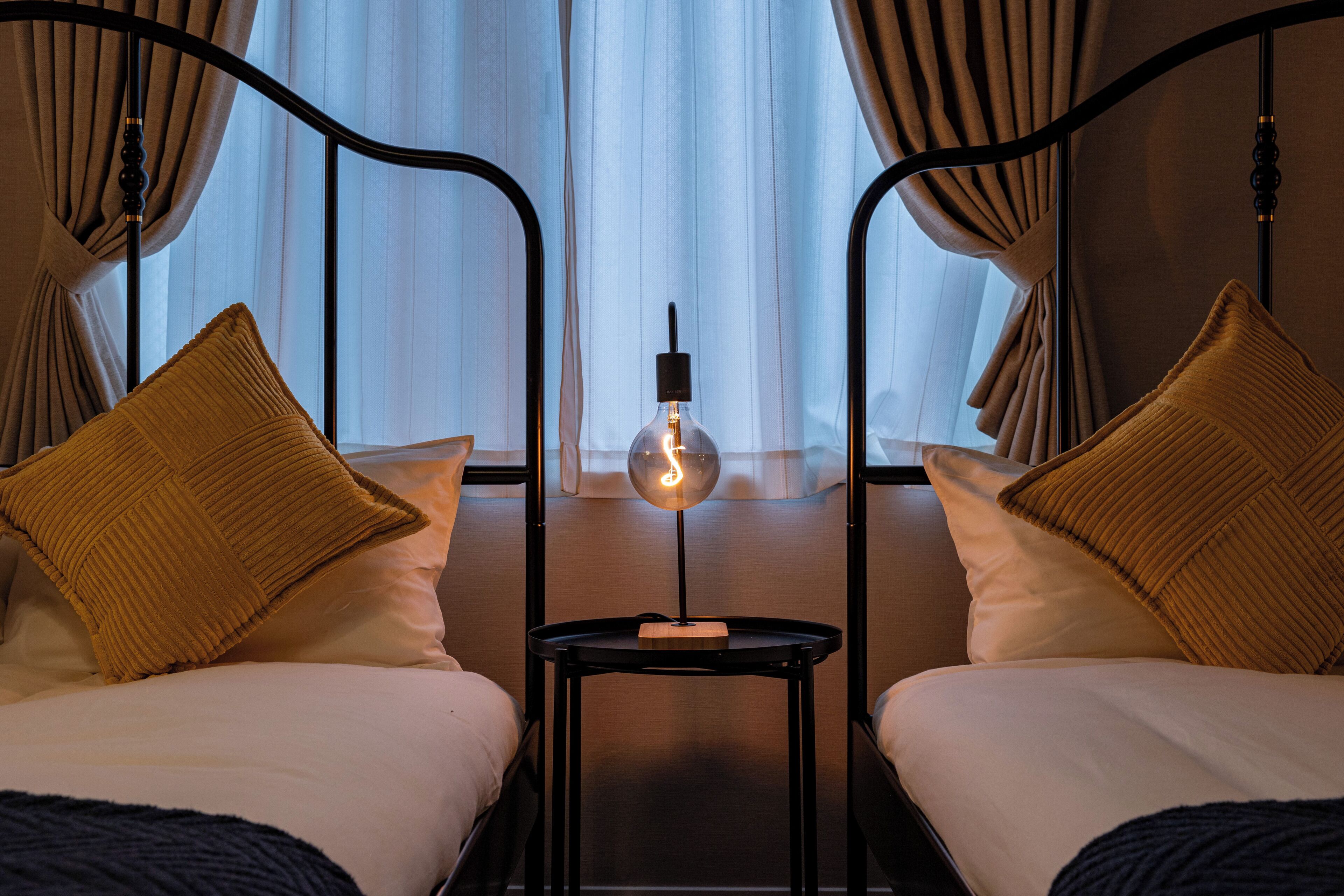 Une chambre d’hôtel avec deux lits, une table de chevet avec une lampe et une vue sur l’extérieur à travers des rideaux transparents.
