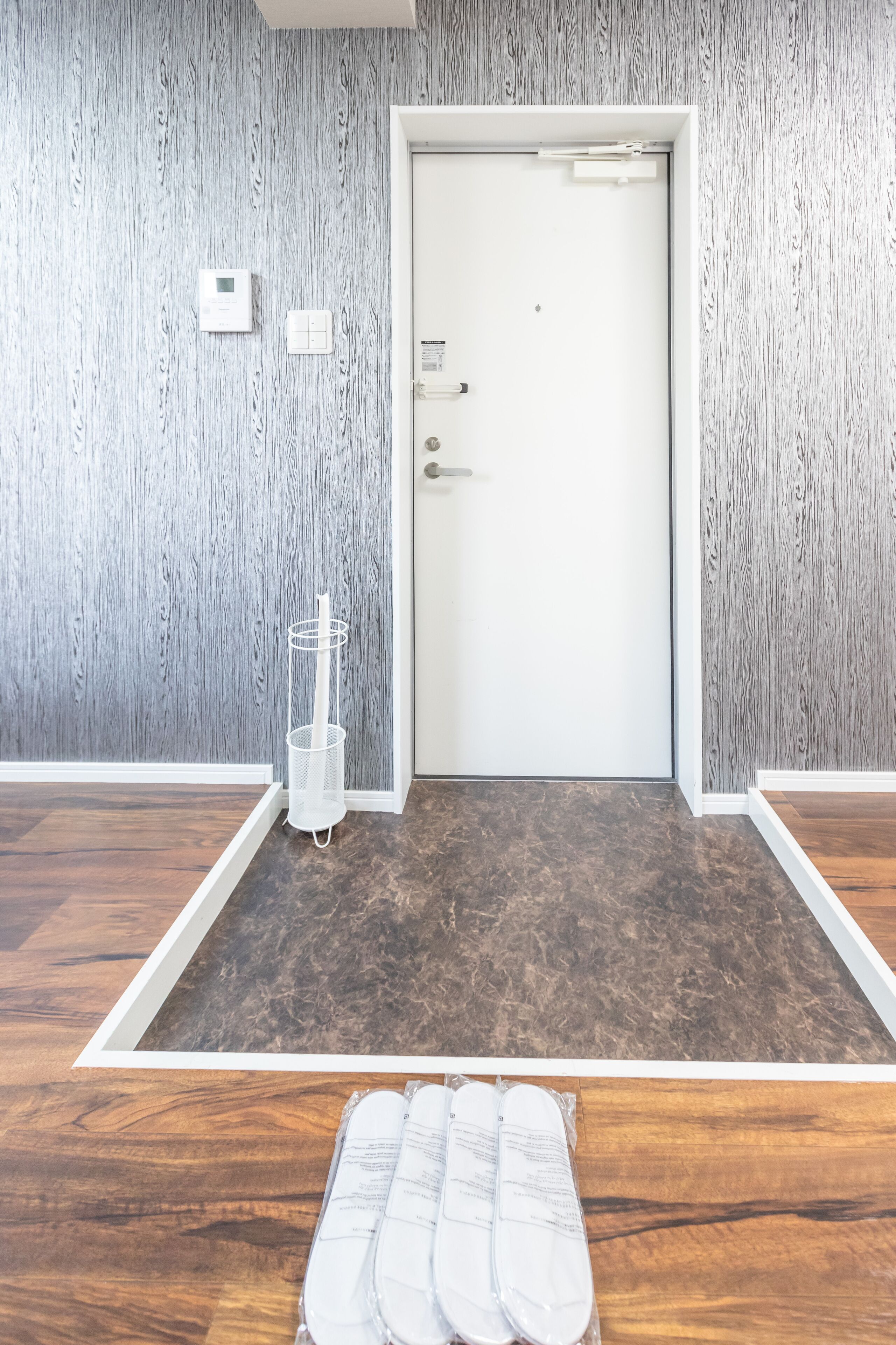 A room with a white door, a white wall with a vertical wood grain pattern, a white floor, and a brown wooden floor.