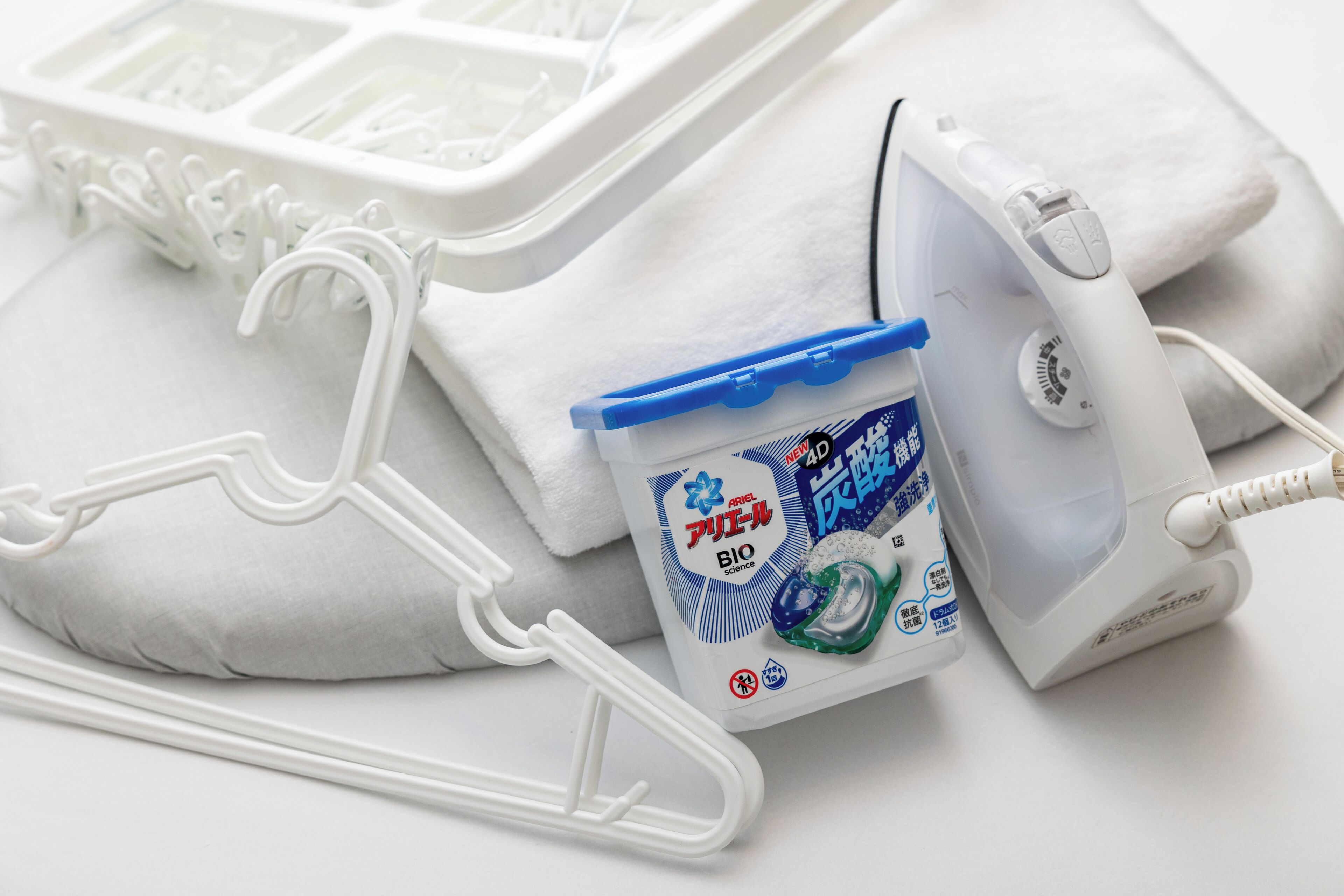 Un cintre en plastique blanc, un fer à repasser blanc et un bidon de lessive Ariel Bio.