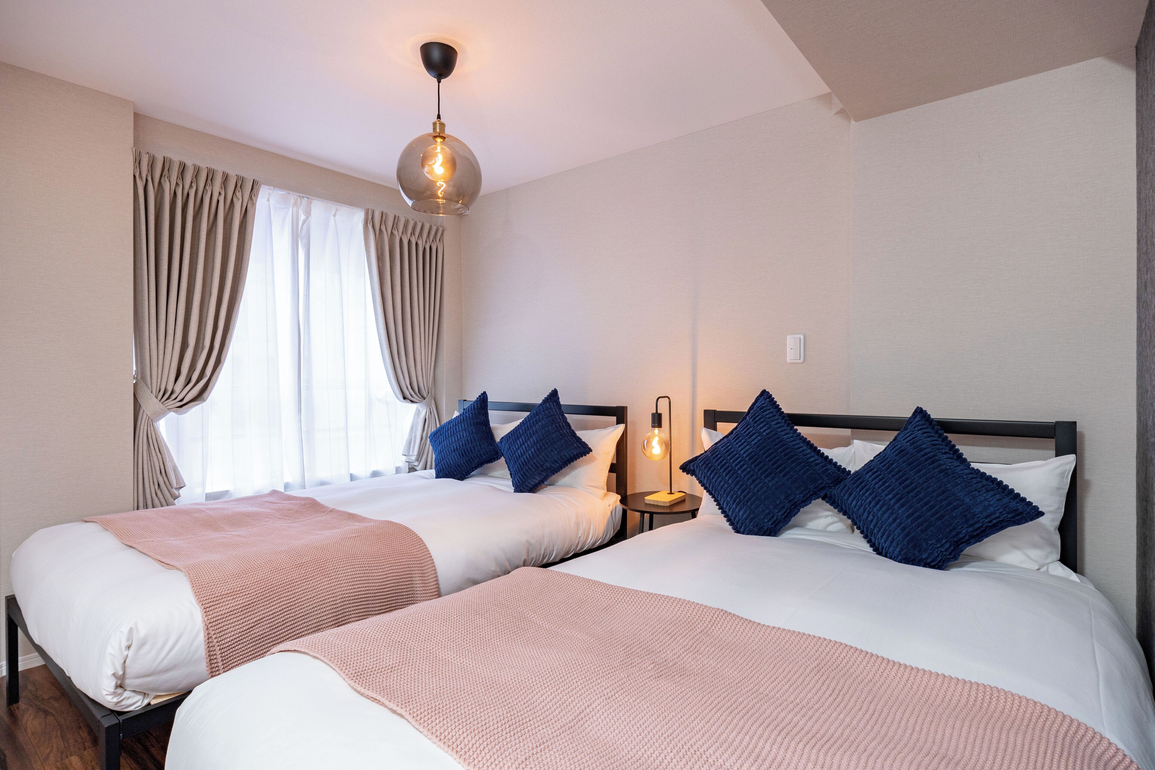 A hotel room with two beds, beige curtains, a hanging light fixture, and a bedside table with a lamp.