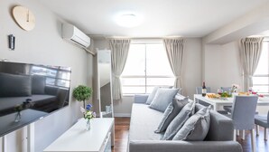 A modern living room with a flat-screen TV, grey sofa, dining table with chairs, and a small side table with a vase of flowers. - Arita Nipponbashi-nishi (Osaka)