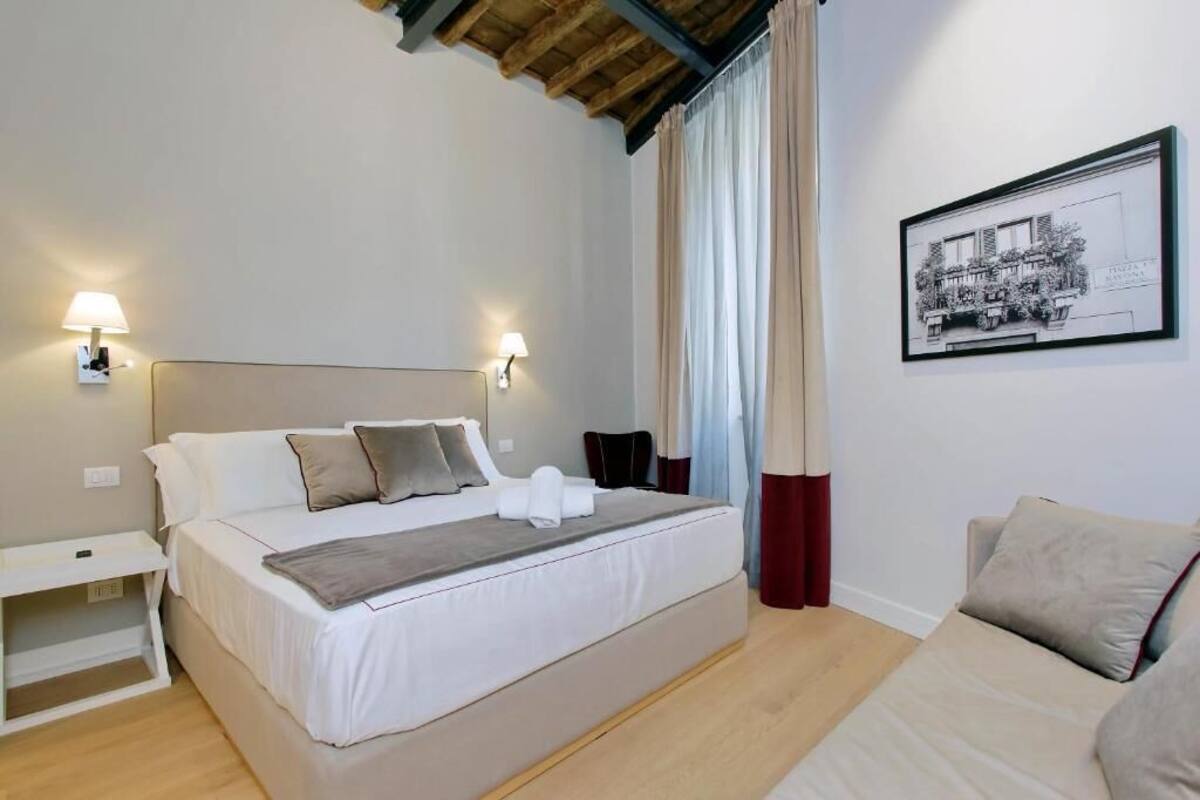 A bedroom with a bed, bedside tables, a nightstand, a headboard, a wall-mounted lamp, a window with curtains, and a framed picture on the wall.
