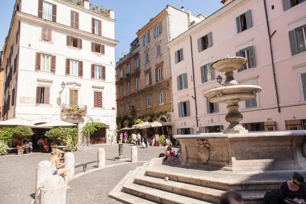 Une place publique agrémentée d’une fontaine, entourée de bâtiments historiques dotés de balcons et d’espaces de détente en plein air.