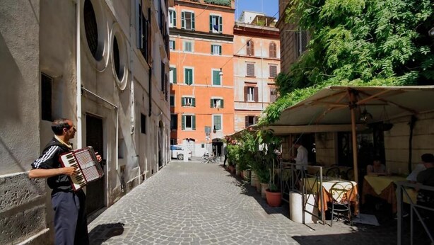 A cobblestone street with buildings on either side, a musician playing an accordion, and outdoor seating at a cafe. - Monti Panisperna Suites (Rome)