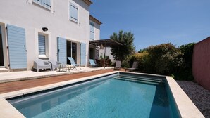 Una piscina con plataforma de madera, zona de estar cubierta y un edificio blanco con contraventanas azules.