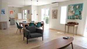 Amplia sala de estar con una mesa de centro de madera, un sillón gris y una mesa de comedor con sillas. Se observa una cocina al fondo, un cuadro en la pared y una ventana que proporciona luz natural.