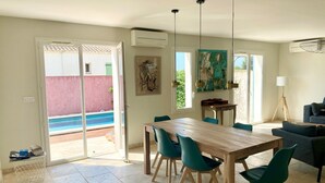 Un comedor con mesa de madera y sillas color turquesa, un cuadro en la pared y vista a una piscina a través de puertas correderas.