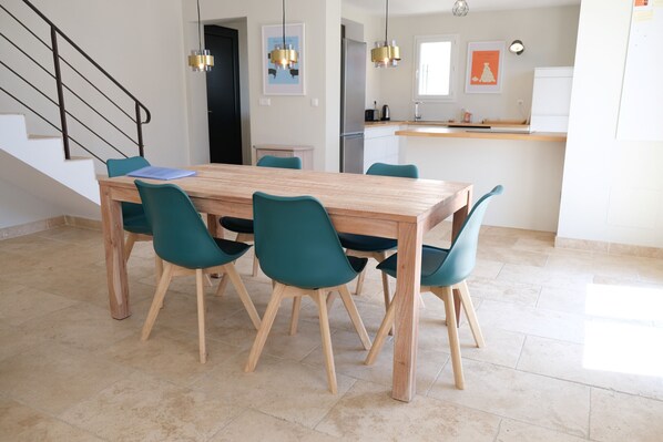 Un área de comedor moderna con una mesa de madera y sillas color turquesa, una cocina al fondo y una escalera con barandales negros.