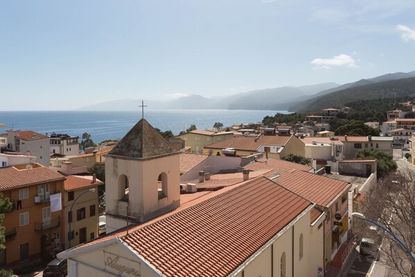 A coastal town with terracotta-roofed buildings, a prominent church with a bell tower, and a view of the sea and mountains in the background.