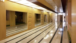 Pasillo de hotel con varias habitaciones, cada una con puerta y pequeña ventana. El pasillo tiene suelo de baldosas y está bien iluminado con luces del techo.