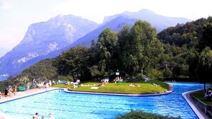 Una piscina con montañas al fondo, rodeada de exuberante vegetación y gente descansando sobre el césped.