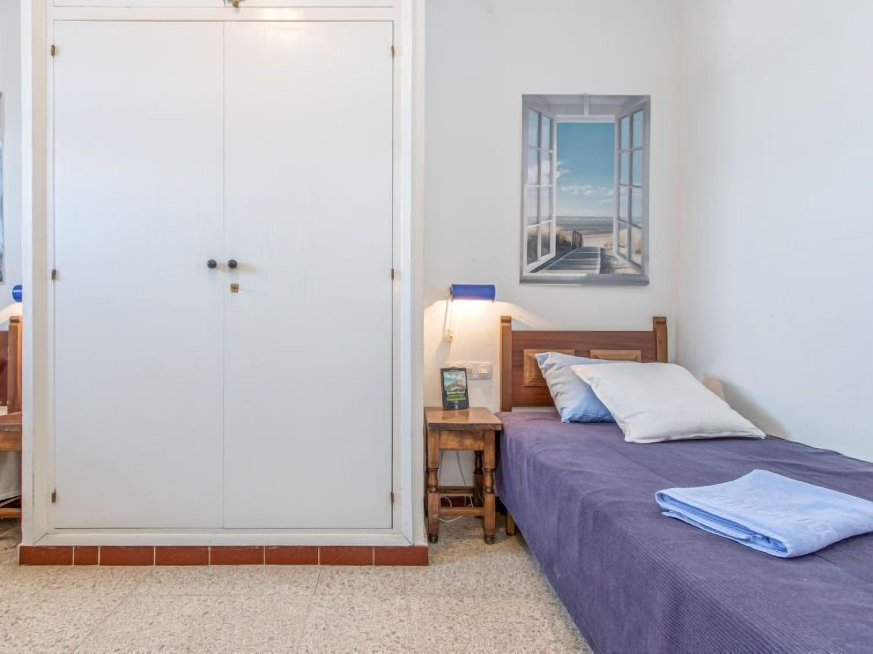 A bedroom with a bed, a nightstand, a lamp, a wardrobe, and a painting on the wall.