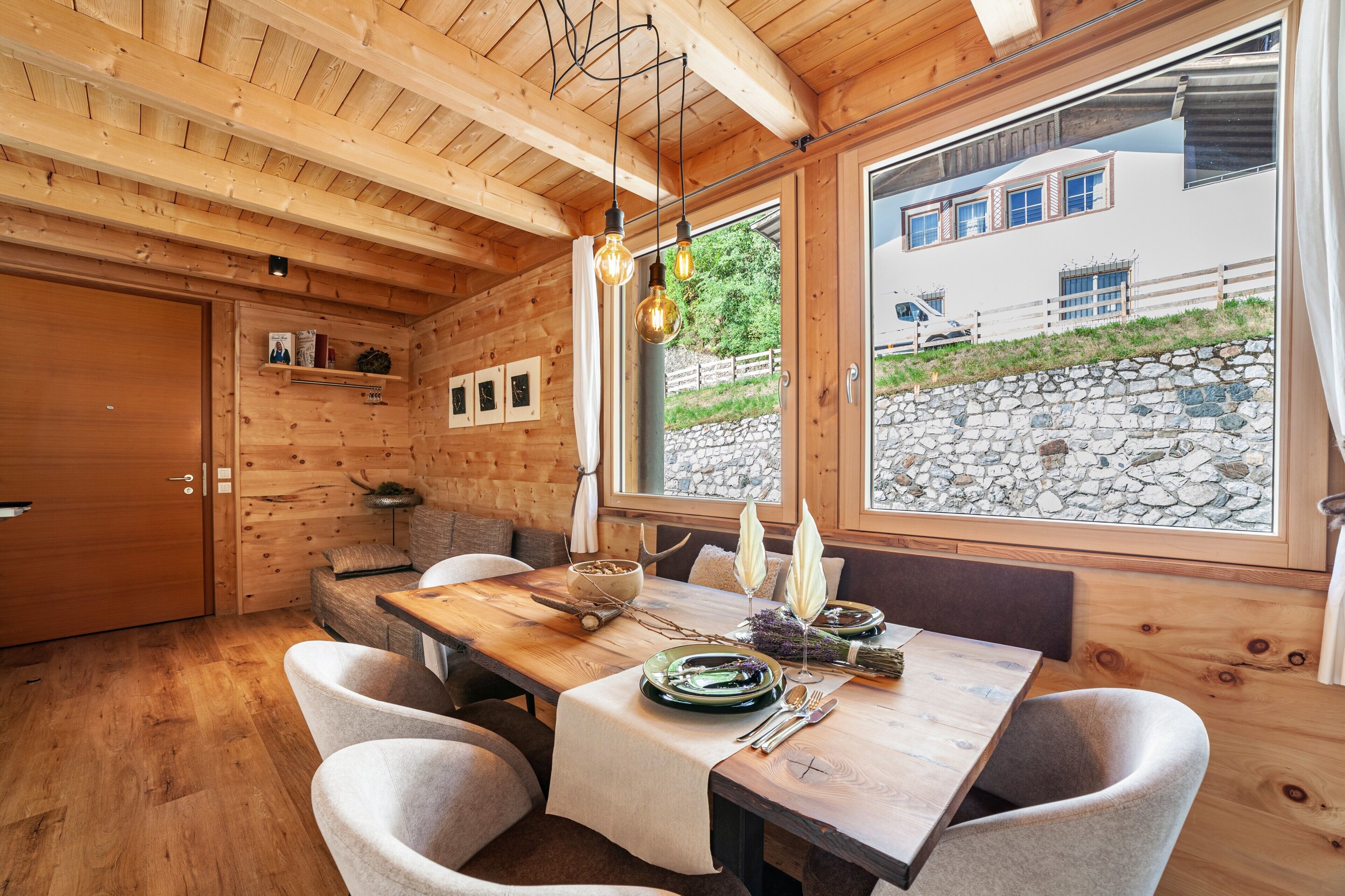 Un intérieur en bois avec une table à manger dressée pour quatre personnes, un banc et une vue sur un mur de pierre à l’extérieur.