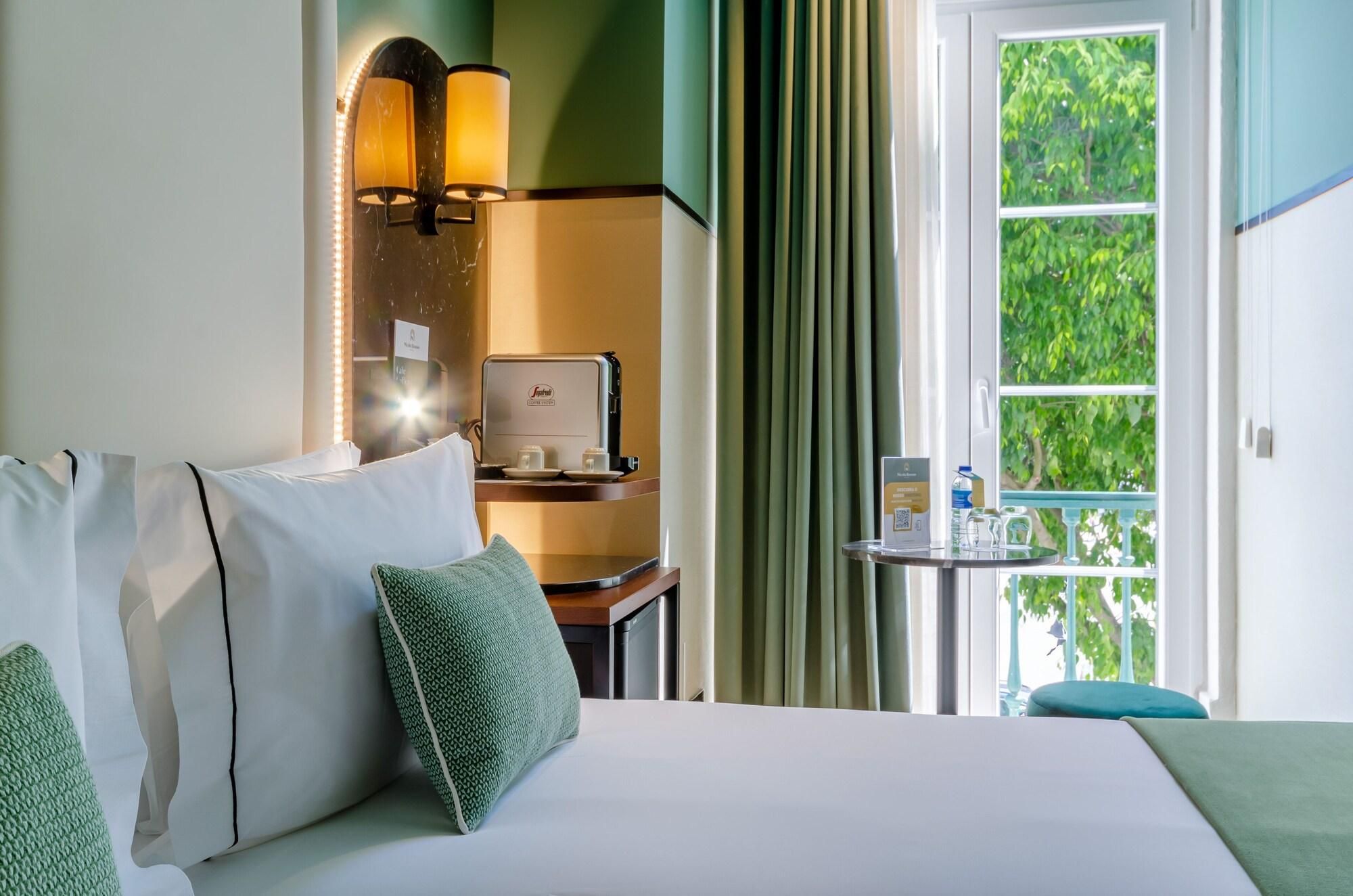 Une chambre d’hôtel avec un lit, des oreillers, un bureau avec un ordinateur portable, une petite table avec une bouteille et un verre, et une fenêtre avec des rideaux.