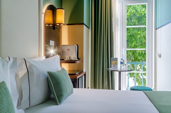 A hotel room with a bed, pillows, a desk with a laptop, a small table with a bottle and a glass, and a window with curtains.