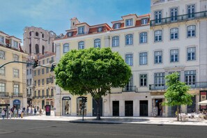 Une scène de rue avec un grand arbre au premier plan, des immeubles à plusieurs étages et quelques piétons.