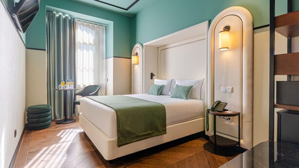 Une chambre d’hôtel moderne avec un grand lit, un bureau et une chaise. On y trouve une fenêtre avec des rideaux, un téléphone fixé au mur et une petite table avec une lampe.