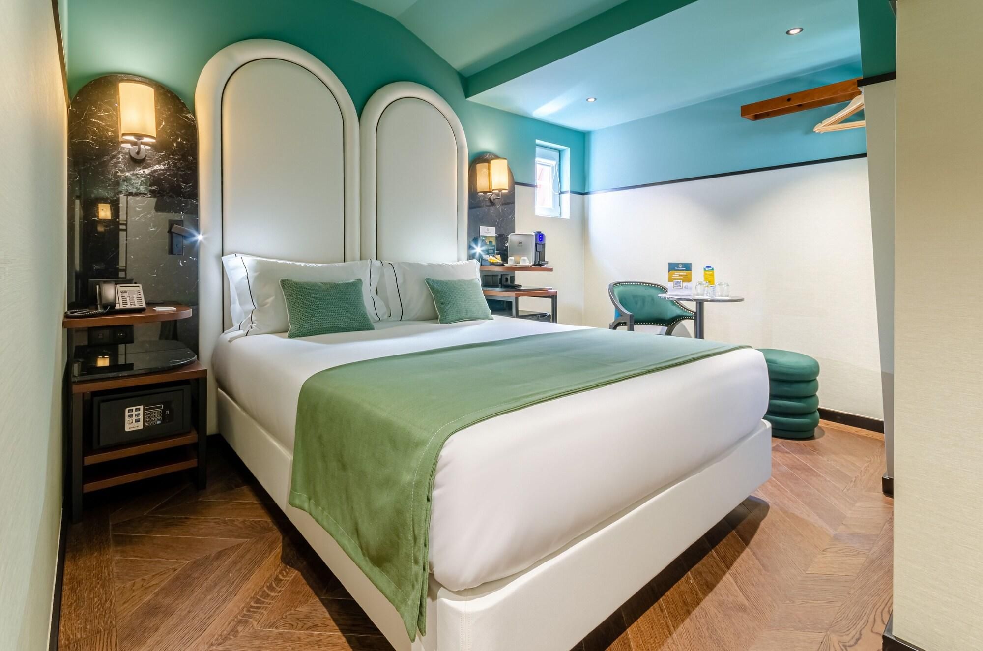 Una habitación de hotel moderna con una cama grande, un escritorio con silla, una mesita pequeña y un reposapiés verde.