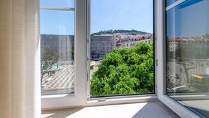 Une chambre d’hôtel dotée d’une grande fenêtre donnant sur un paysage urbain historique, avec ses bâtiments et sa fontaine.