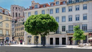 Une scène de rue avec un grand arbre au premier plan, des immeubles à plusieurs étages et quelques piétons.