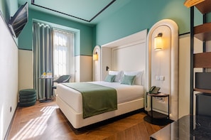 A modern hotel room with a large bed, a desk, and a chair. There is a window with curtains, a wall-mounted phone, and a small table with a lamp.