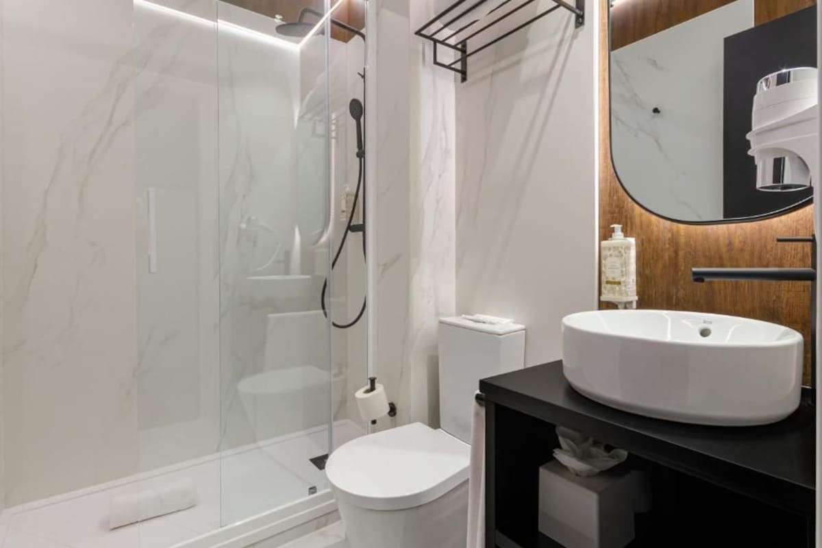 Un baño moderno con ducha de vidrio, inodoro blanco y tocador negro con un espejo grande.