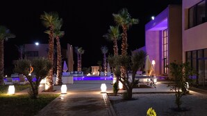 A nighttime outdoor area with a paved walkway, palm trees, and illuminated seating areas. - SUMAHAN SUITES & SPA (Tameslouht)