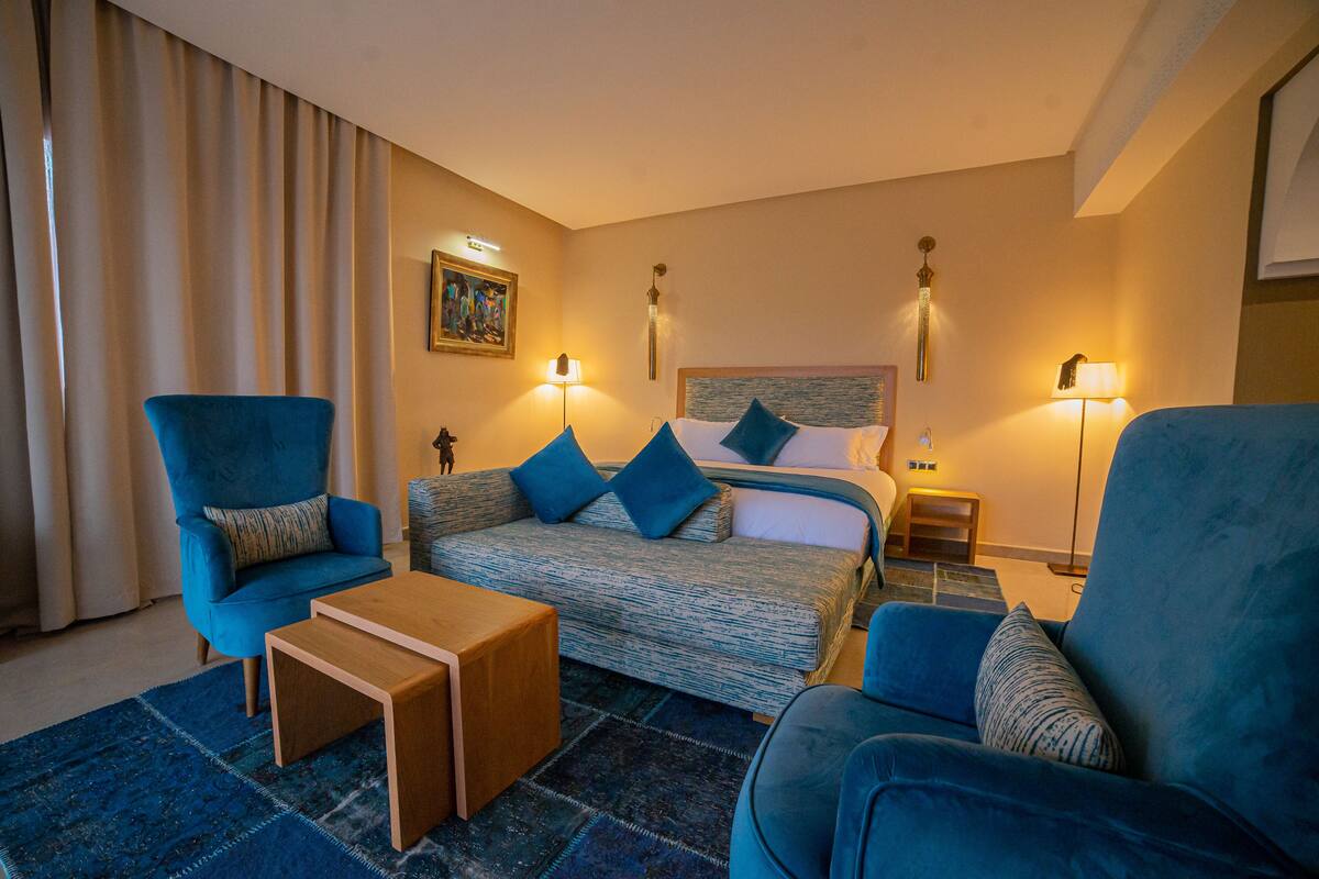 A hotel room with a bed, two blue armchairs, a wooden coffee table, and a patterned rug.