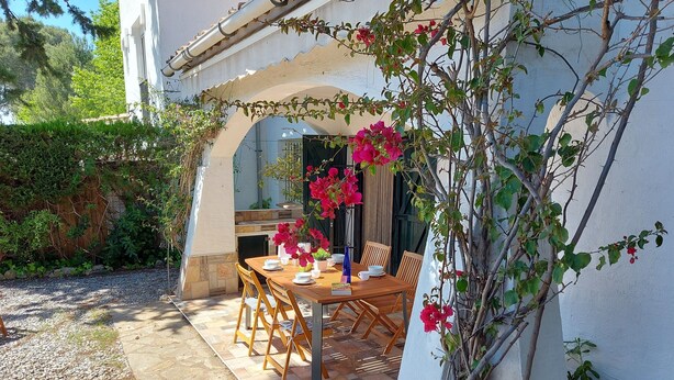 An outdoor dining area with a wooden table and chairs, surrounded by a white wall and greenery. - Typical house under the pines with swimming pool and wifi (L'Escala)