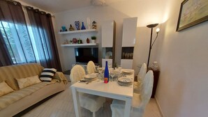A dining area with a white table set for four, surrounded by white chairs. A sofa with striped cushions is in the background, and a TV is mounted on the wall. - Typical house under the pines with swimming pool and wifi (L'Escala)