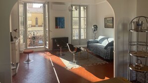 A room with a large arched doorway, a radiator, a sofa, a chair, a small table, and a metal shelf with glasses and a pitcher. - Center, charm, comfort, Clim, shops, restaurants, transport, outings, pkg. (Toulon)