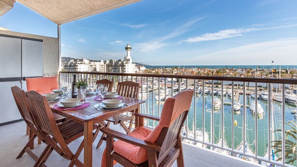 A balcony with a table set for dining, overlooking a marina with boats and a coastal town in the background.